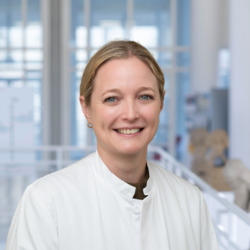 Porträt einer Mitarbeitenden der Hautklinik am Universitätsklinikum Essen mit zurückgebundenem blondem Haar und weißem Kittel, aufgenommen im Klinikgebäude vor lichtdurchfluteter Glasarchitektur