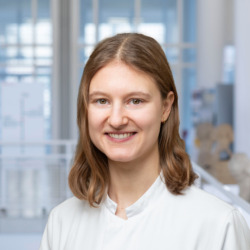 Nahaufnahme von Sarah Kochendörfer, einer Mitarbeitenden der Hautklinik am Universitätsklinikum Essen mit schulterlangem, braunem Haar und weißem Kittel, aufgenommen vor moderner Glasarchitektur im Klinikgebäude